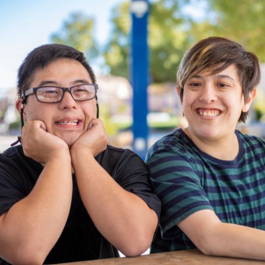 Photo of two boys with developmental disabilities smiling. One is wearing a black shirt and has his hands under his shirt. the other is wearing a blue and green striped shirt and smiling at the camera.