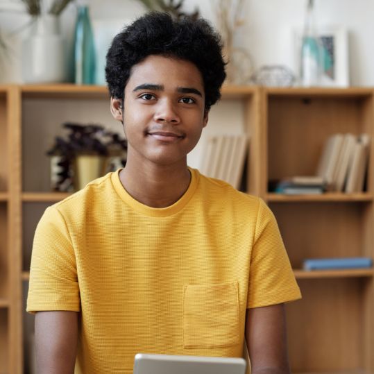 photo of a youth with a tablet. He is wearing a yellow shirt and smiling.